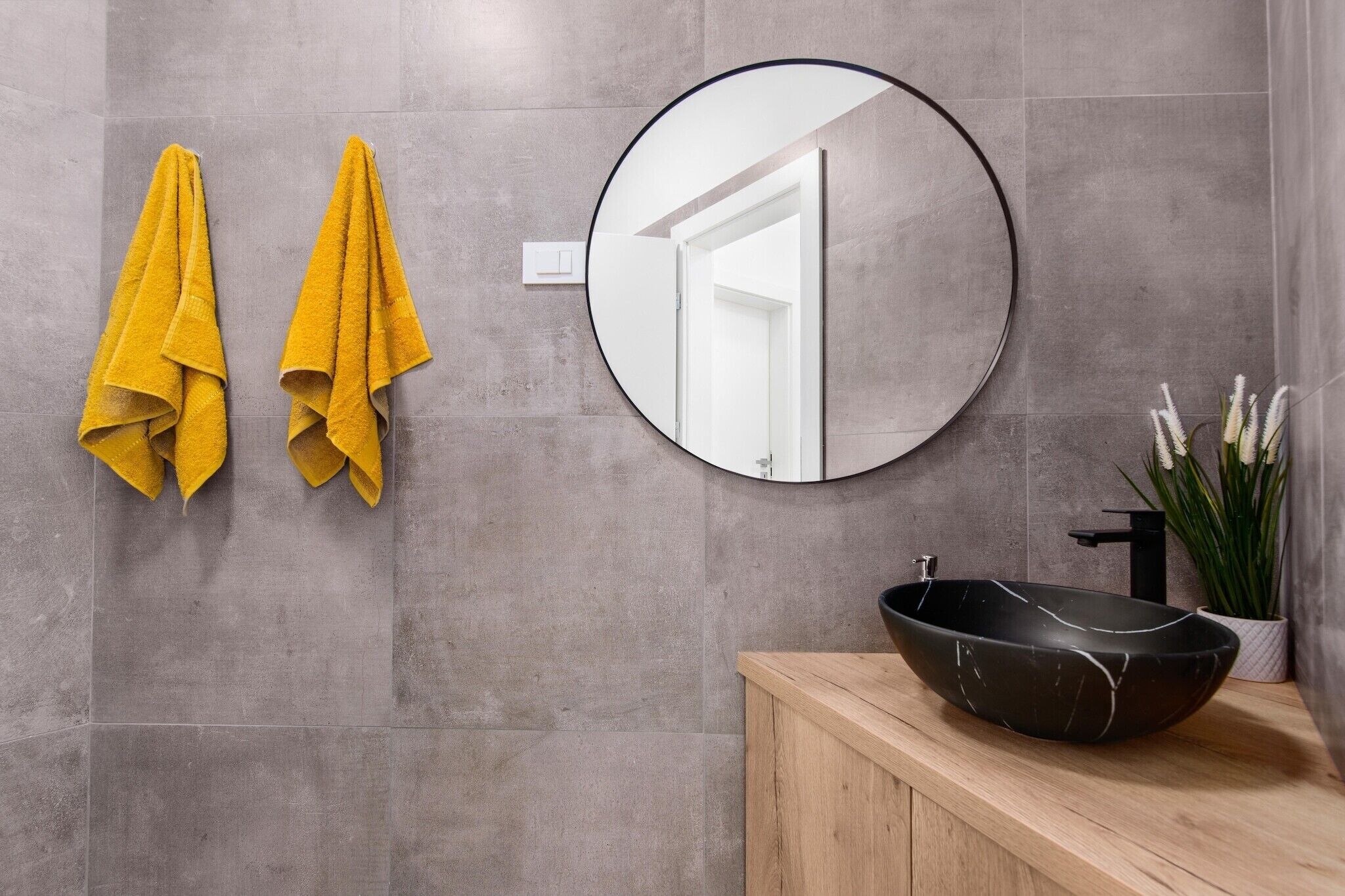 A bathroom with grey tiles, a round mirror, two yellow towels, a black sink, and a potted plant.