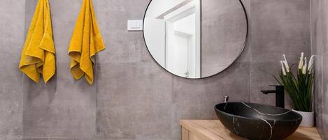 A bathroom with grey tiles, a round mirror, two yellow towels, a black sink, and a potted plant.