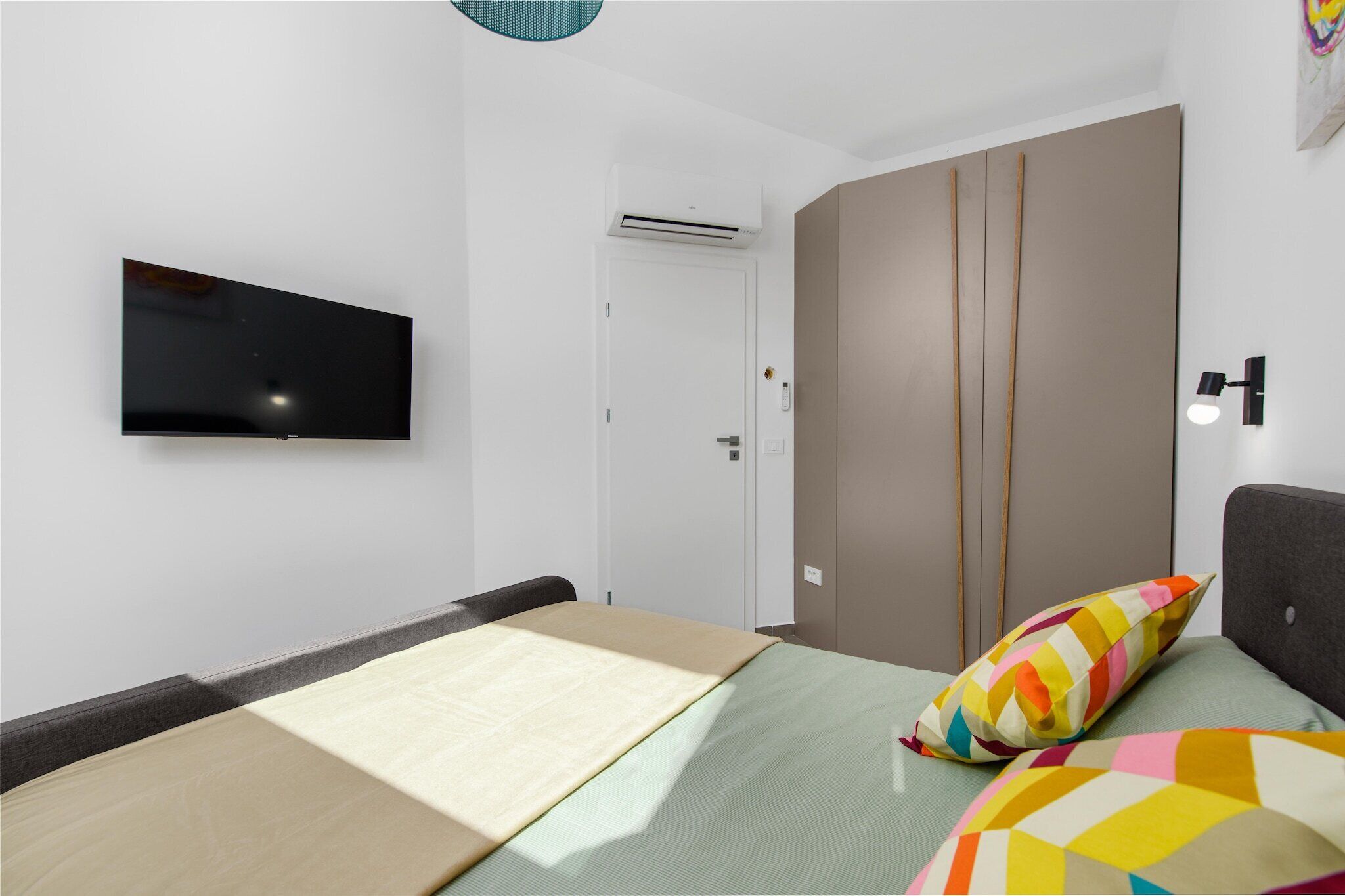 Une chambre à coucher moderne, avec un lit, un téléviseur fixé au mur, une armoire et une table de chevet avec une lampe.
