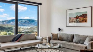 A modern living room with a large window offering a mountain view, a framed landscape painting, and a glass coffee table. - Electric Pass Lodge 604 (Snowmass)