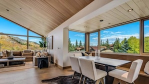 Un salon moderne doté d’une grande table à manger, de sièges confortables et de larges fenêtres offrant une vue panoramique sur les arbres et les montagnes.