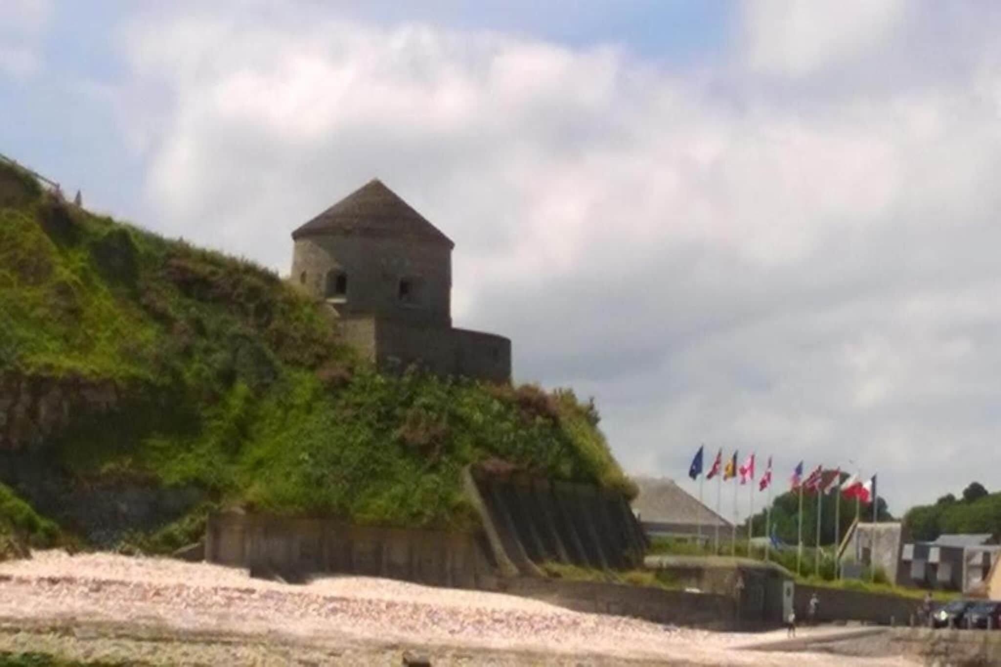 A stone building with a conical roof on a grassy hill, flags in the background, and a clear sky.