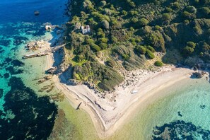 An aerial view of a coastal area with clear blue waters, a sandy beach, and dense green vegetation.