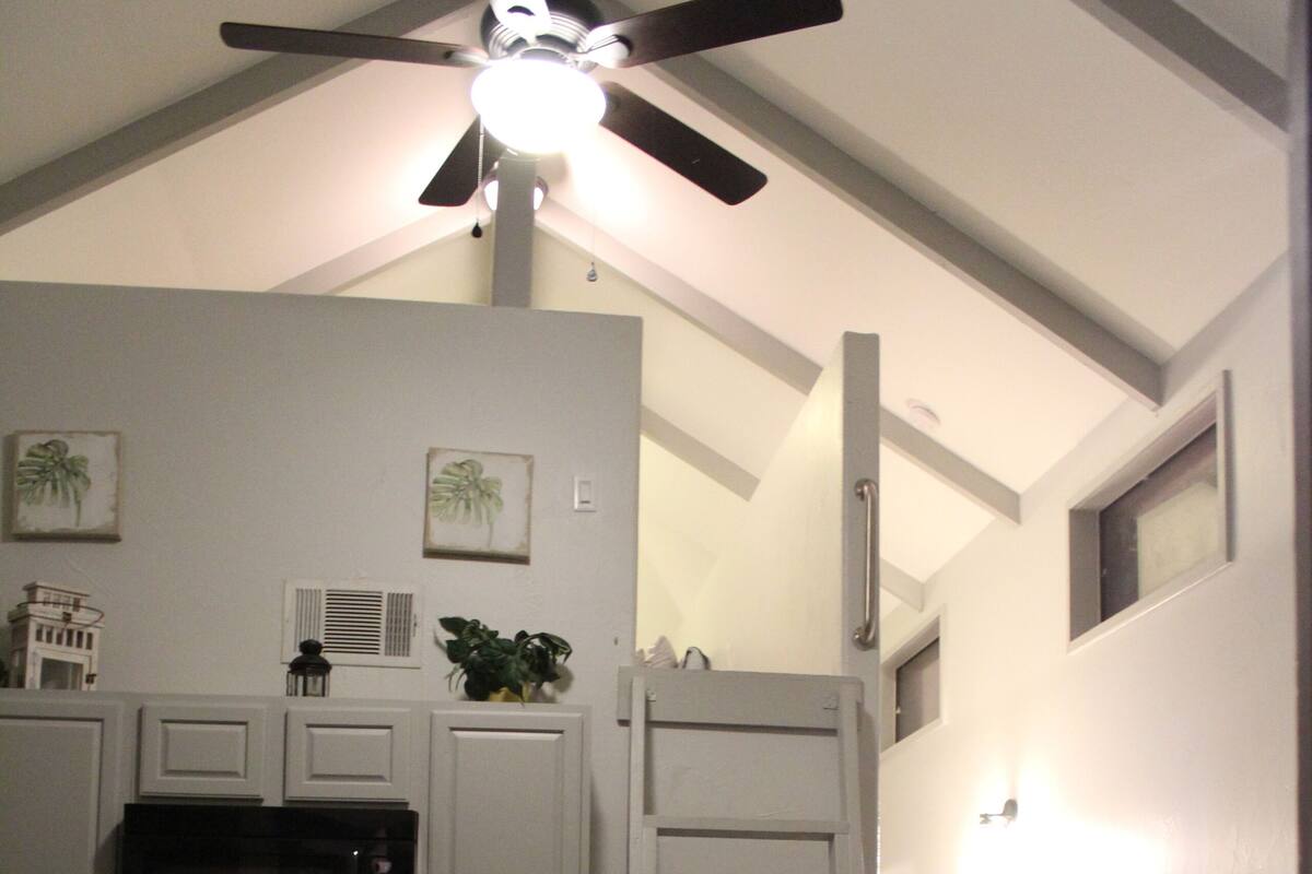 A compact kitchen with white cabinets, a microwave, and a stove. There is a dining table with chairs and a ceiling fan. A ladder is propped against the wall, and a couch is visible in the corner.