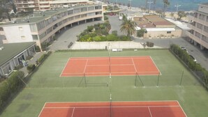 Ein Tennisplatz mit angrenzendem Gehweg, benachbarten Gebäuden und Blick aufs Meer.