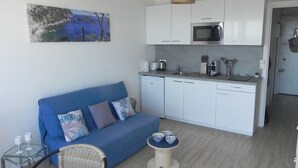 Ein gemütlicher Wohnbereich mit Küchenzeile, einem blauen Sofa mit gemusterten Kissen, einem Korbstuhl und einem kleinen Tisch mit dekorativen Schalen.