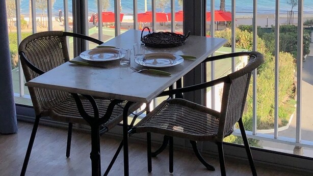 Ein Balkon mit Tisch für zwei Personen, mit Blick auf einen Strand mit roten Sonnenschirmen und Palmen.