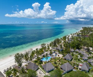 Luchtbeeld van een resort met rieten huisjes, een zwembad en een strand.