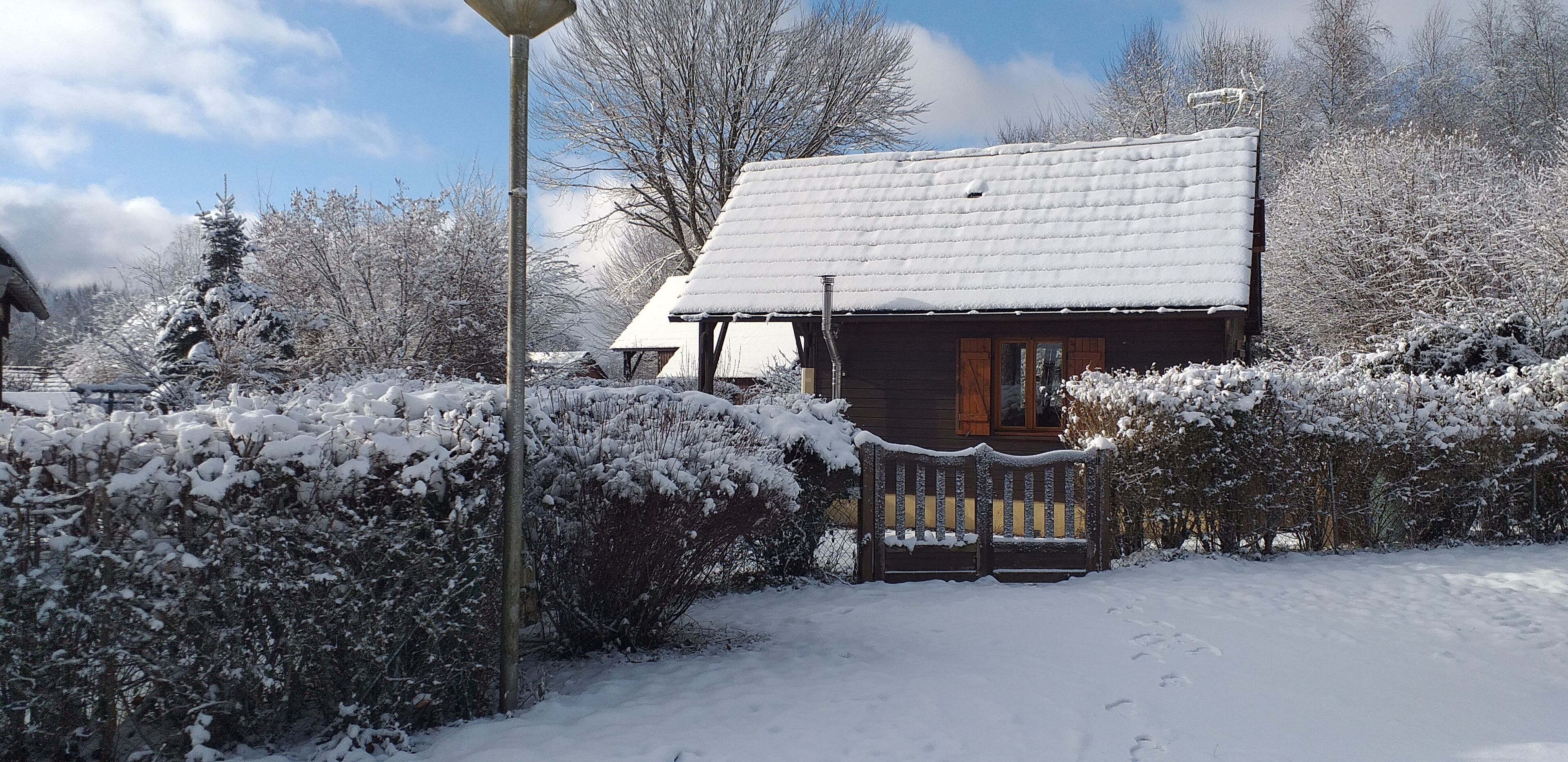 A wooden cabin with a snow-covered roof, surrounded by snow-laden bushes and trees.