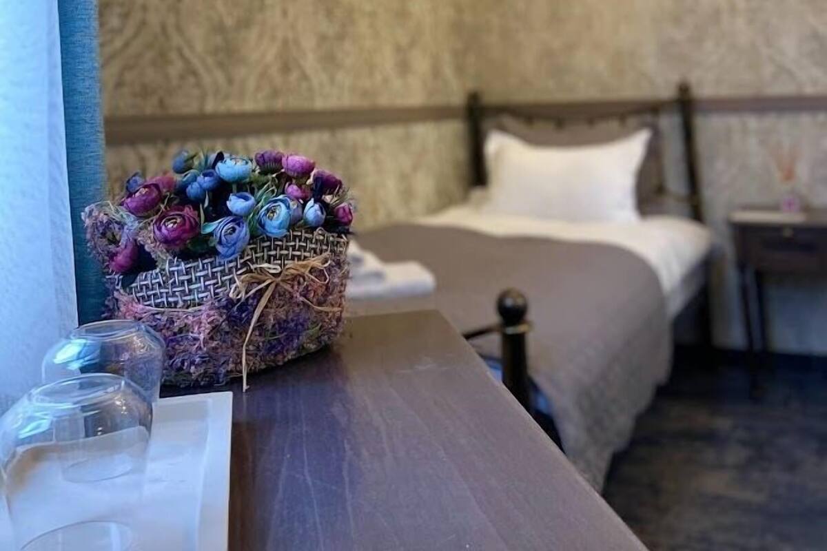 Une chambre d’hôtel avec un papier peint à motifs, un lit recouvert de draps blancs, une table de chevet en bois et une petite table sur laquelle repose un panier en osier contenant des fleurs séchées.