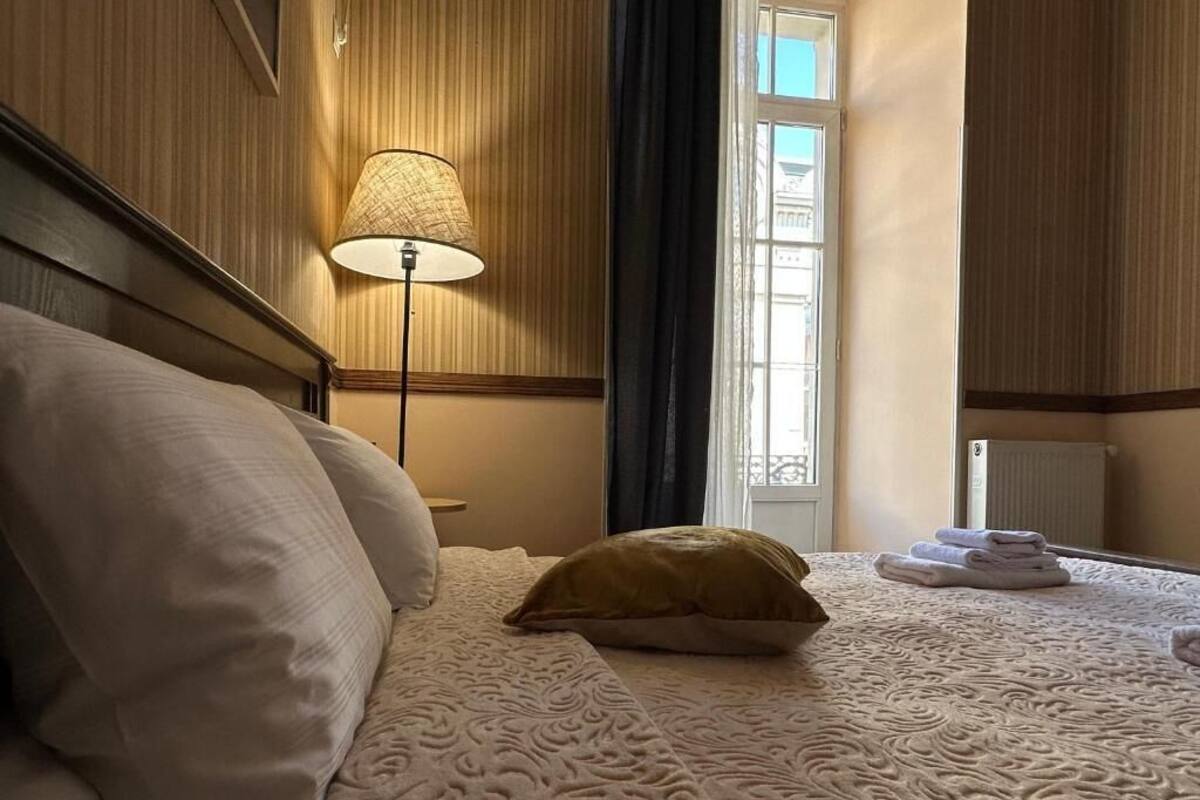Un lit bien fait, recouvert d’une couvre-lit à motifs, une lampe de chevet et une fenêtre avec des rideaux.