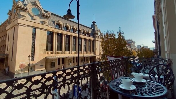 Ein Balkon mit aufwendigen Metallgeländern, einem Tisch mit Tasse und Untertasse sowie ein Gebäude im Hintergrund mit klassischer Architektur.