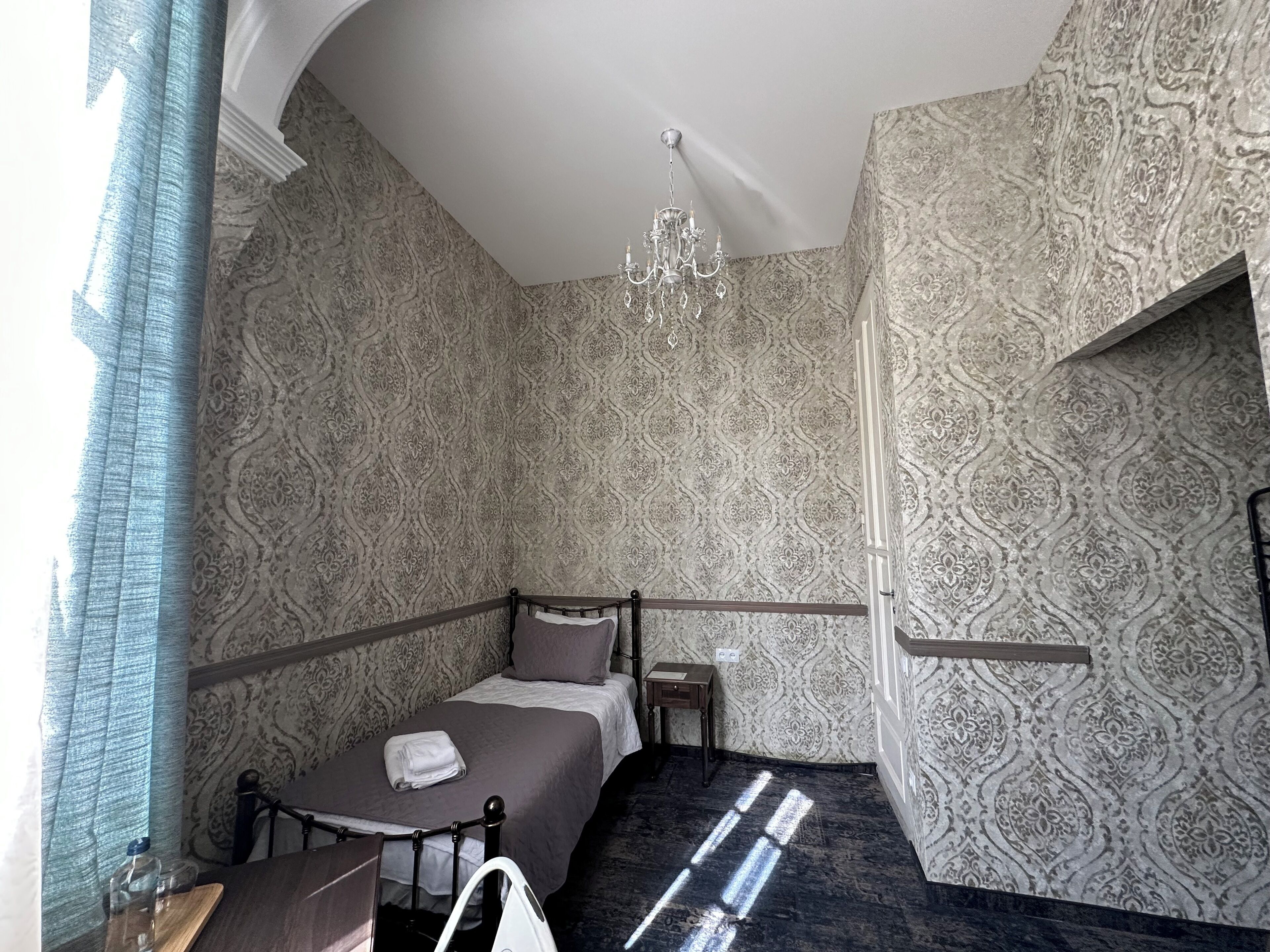 A bedroom with patterned wallpaper, a chandelier, a bed with a brown bedspread, a bedside table, and a nightstand.