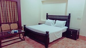 Quarto com cama de madeira, roupa de cama branca, mesa de cabeceira de madeira e uma pequena mesa com uma cadeira.