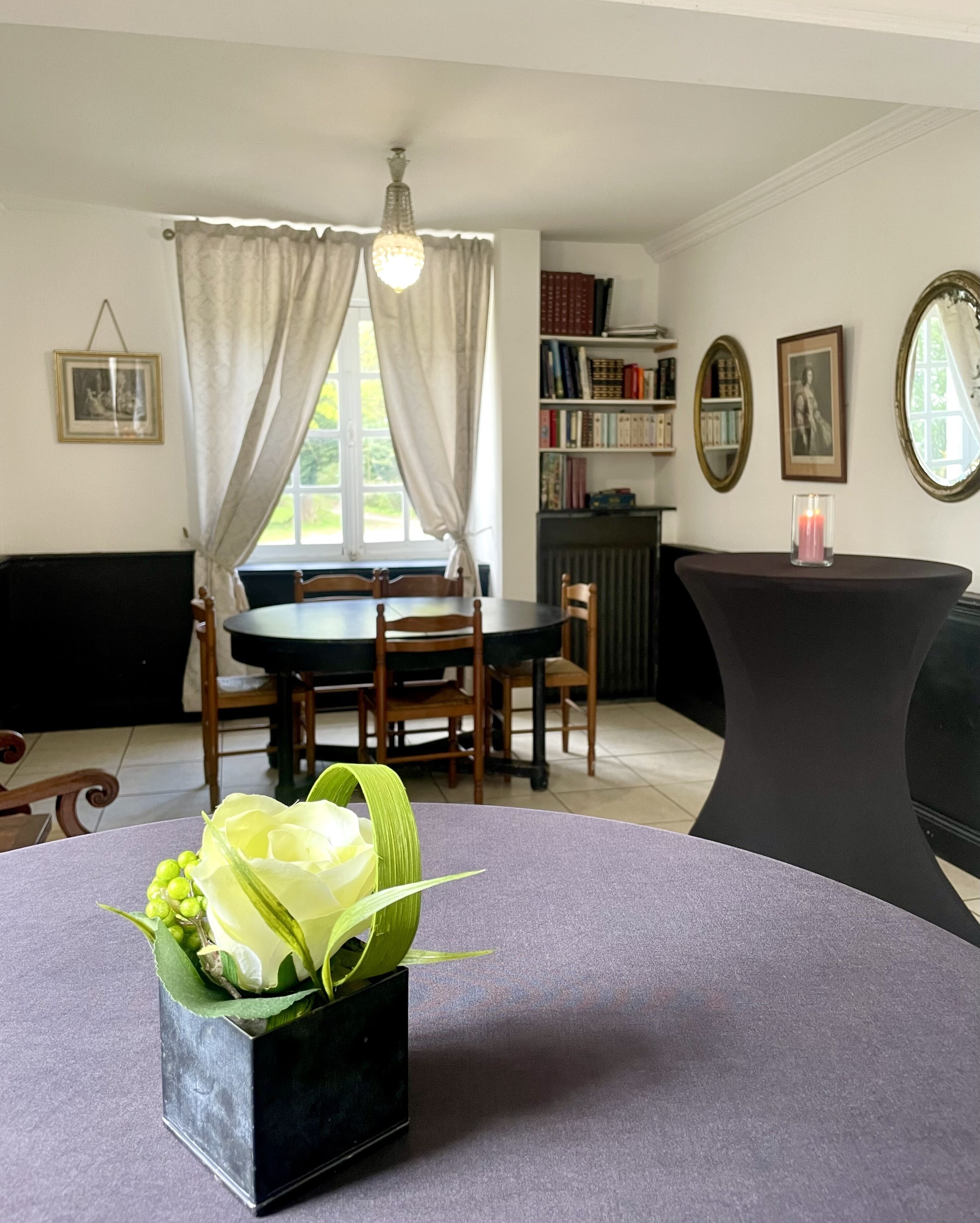 A dining area with a round table, chairs, a bookshelf, and a window with curtains.