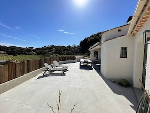 Une terrasse avec des chaises longues et une table, un bâtiment blanc avec un toit en tuiles, et une vue sur des vignobles en arrière-plan.