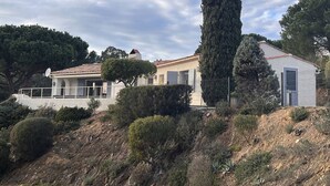 Una villa de estilo mediterráneo con tejado de baldosas, un gran ciprés y un jardín vallado.