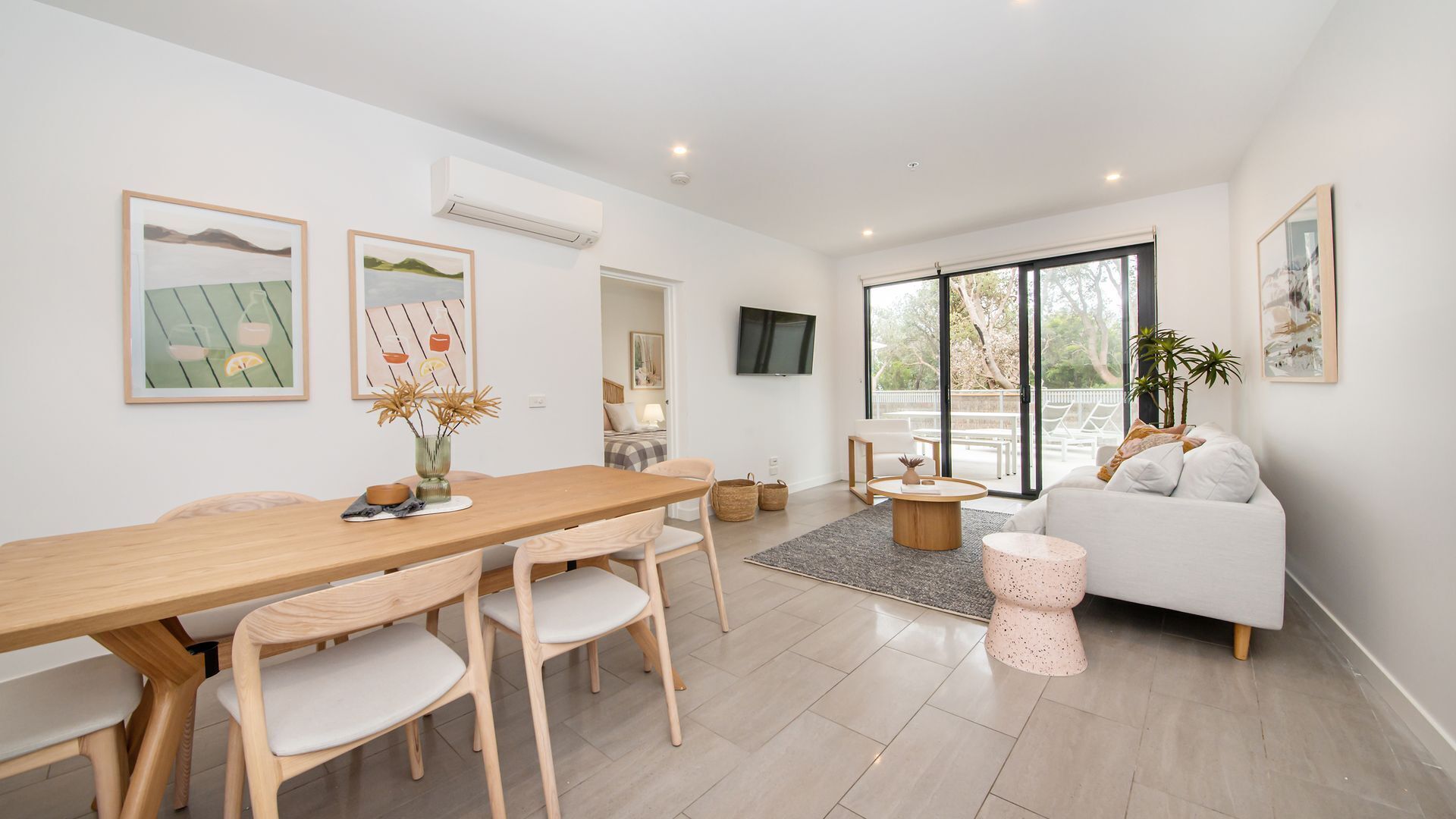 A modern living room with a wooden dining table, white sofa, and a TV mounted on the wall.