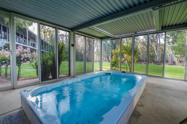 Una piscina techada con paredes de vidrio, rodeada por un jardín y vistas a una casa.