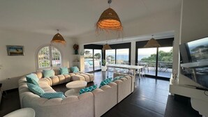 A modern living room with a sectional sofa, a dining table, and large windows offering a view of the sea.