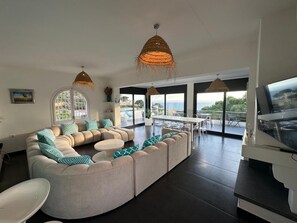 A modern living room with a sectional sofa, a dining table, and large windows offering a view of the sea.