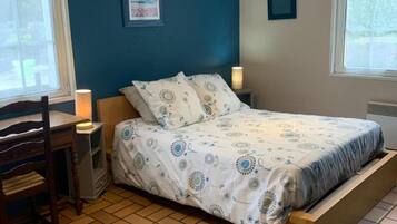 A bedroom with a bed, a desk, a chair, a nightstand with a lamp, and a window.