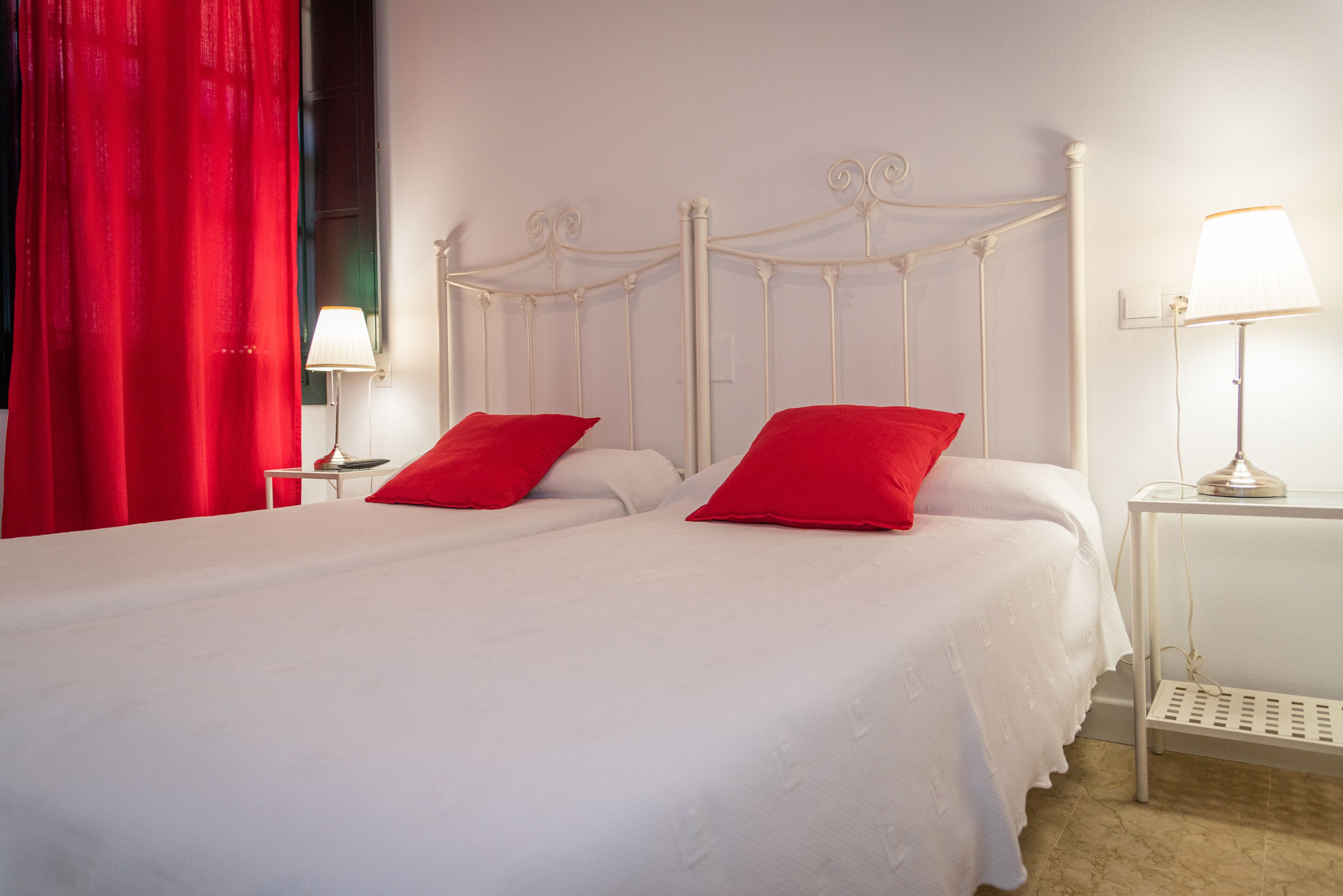 Una cama doble con ropa de cama blanca y dos almohadas rojas, cabecero de metal blanco, mesita de noche con lámpara y cortina roja.