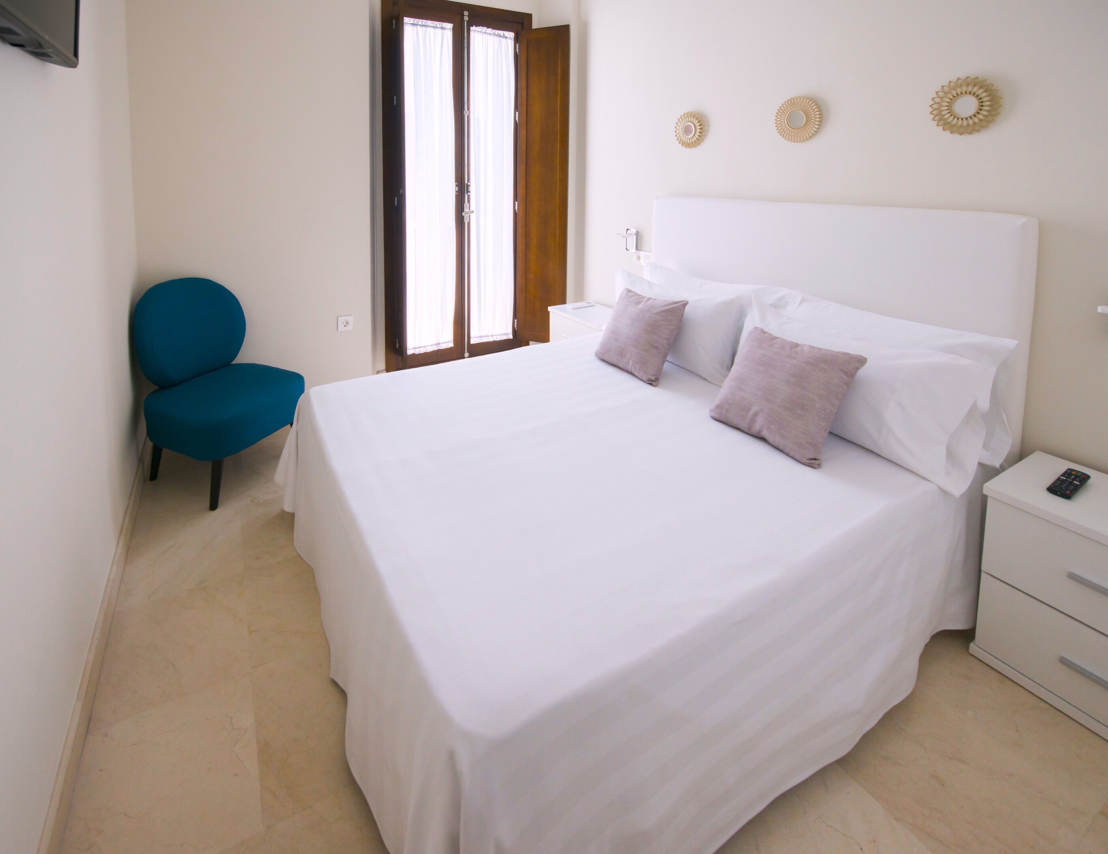 Una cama bien hecha con sábanas blancas y dos almohadas, una silla azul, una mesita de noche con un control remoto y una puerta que da a una vista exterior.