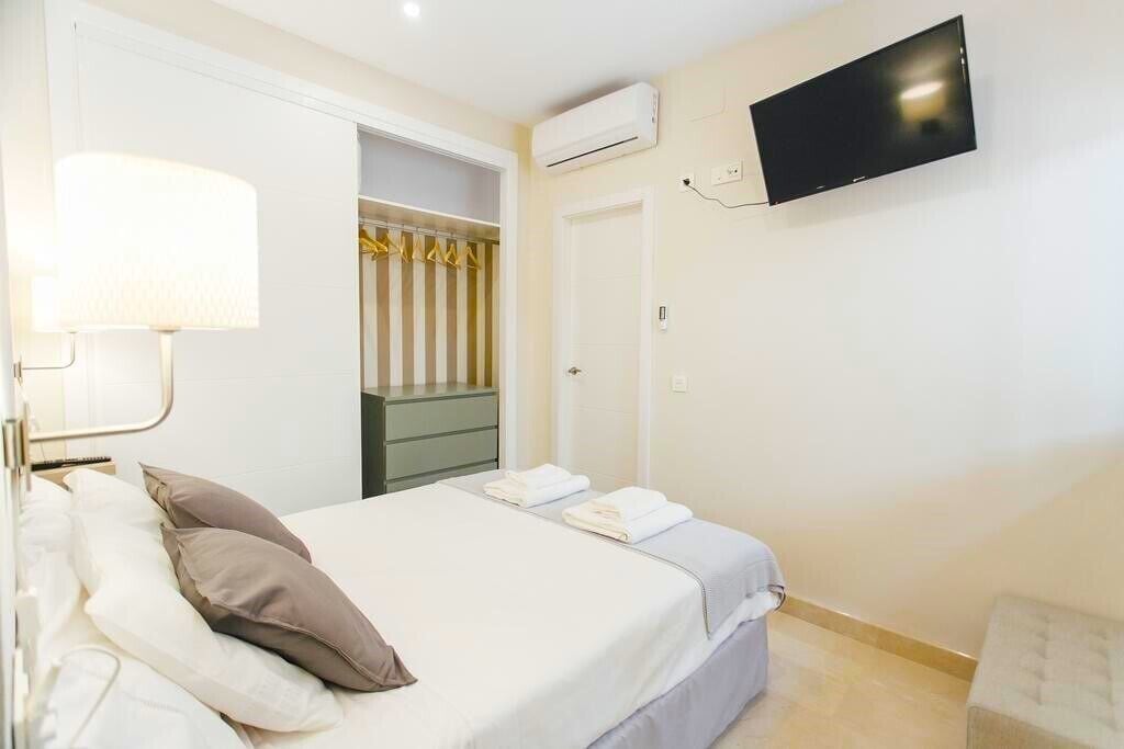 A hotel room with a bed, a TV mounted on the wall, a closet with a striped curtain, and an air conditioning unit.