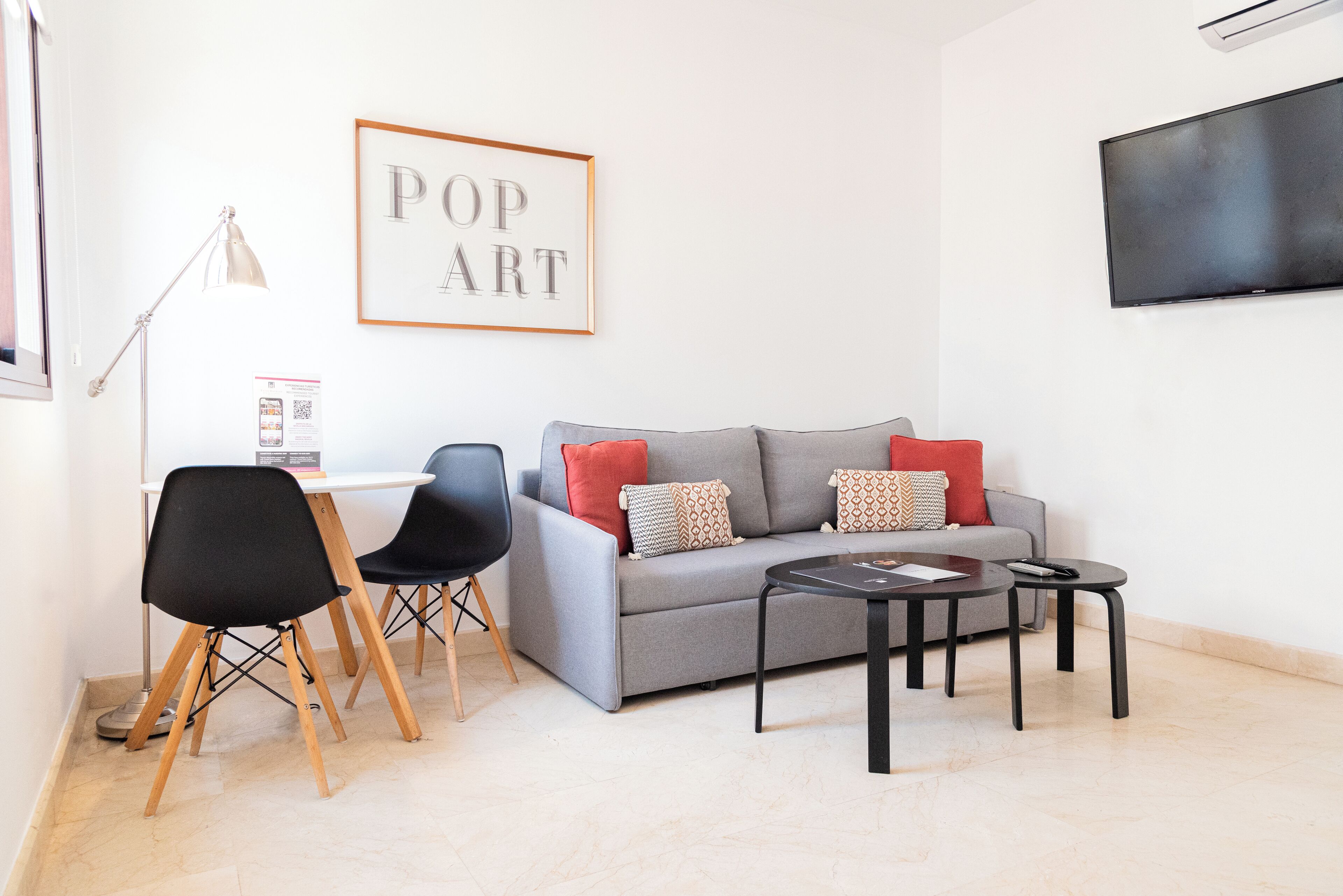 Ein Wohnzimmer mit grauem Sofa, schwarzen Sesseln, einem Couchtisch, einem gerahmten Kunstwerk, einem Fernseher und einem Schreibtisch mit Lampe.
