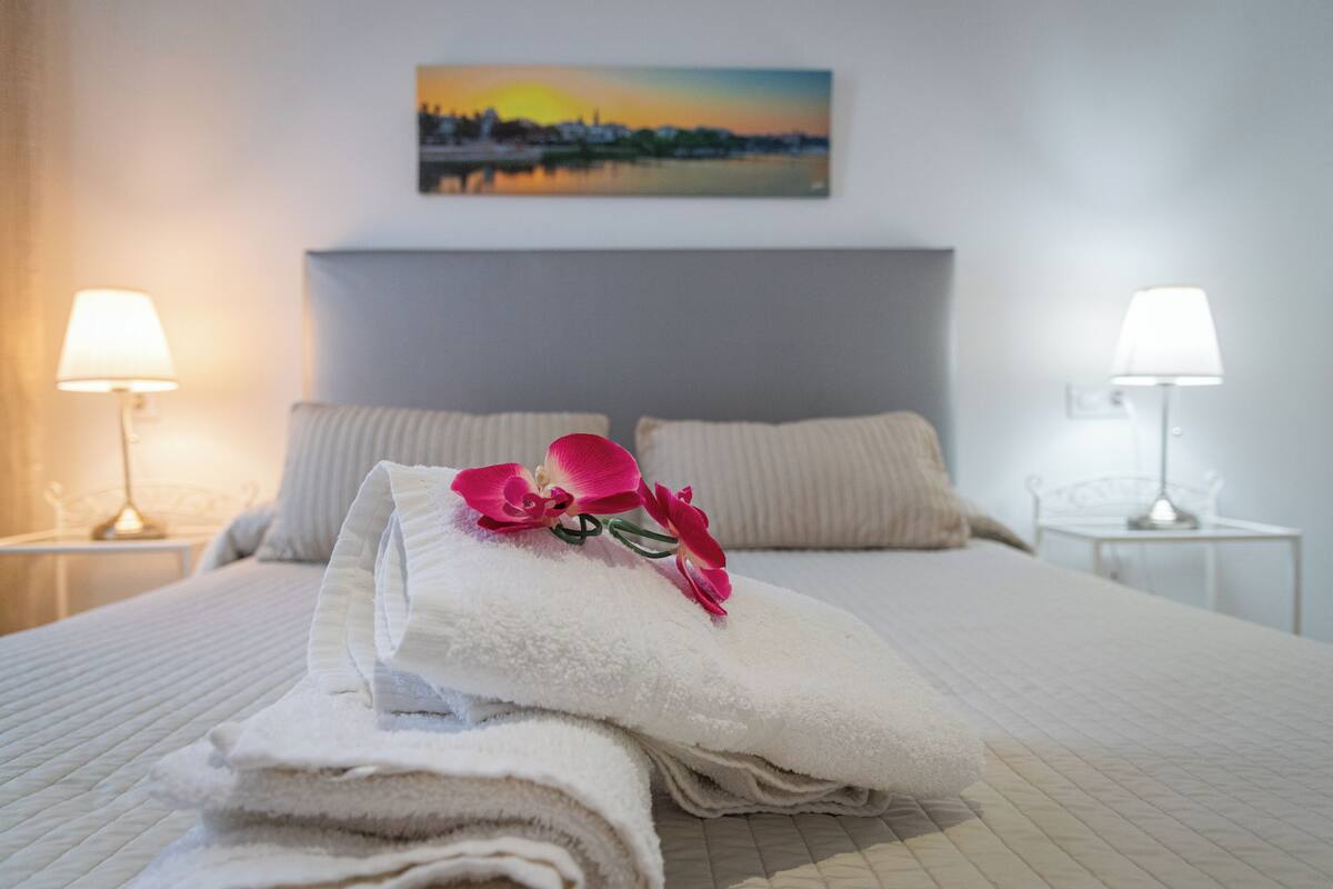 A neatly made bed with folded towels and a decorative orchid, two bedside lamps, and a cityscape painting on the wall.