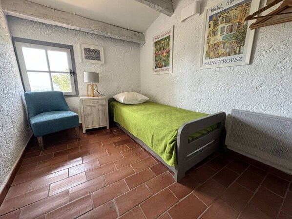 A small, rustic room with a single bed, a chair, a nightstand, and a window.