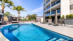 A swimming pool with lounge chairs and a building in the background. - Cove 601 - Luxury living in Forster. (Forster)