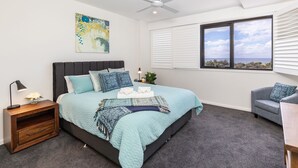 A bedroom with a bed, a nightstand, a chair, a window with blinds, and a ceiling fan. - Cove 601 - Luxury living in Forster. (Forster)
