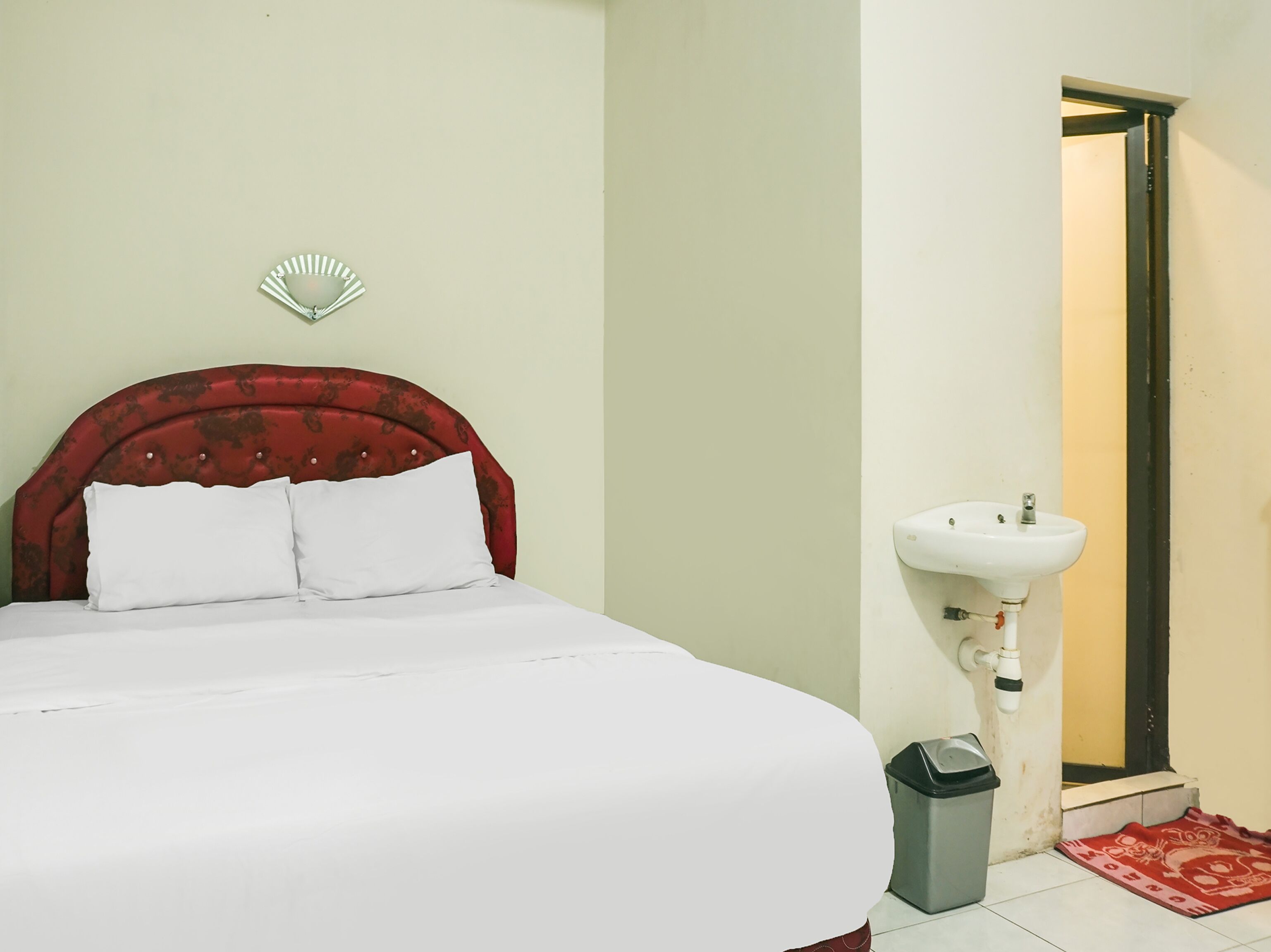 A neatly made bed with white linens, a red upholstered headboard, and a white wall-mounted fan.