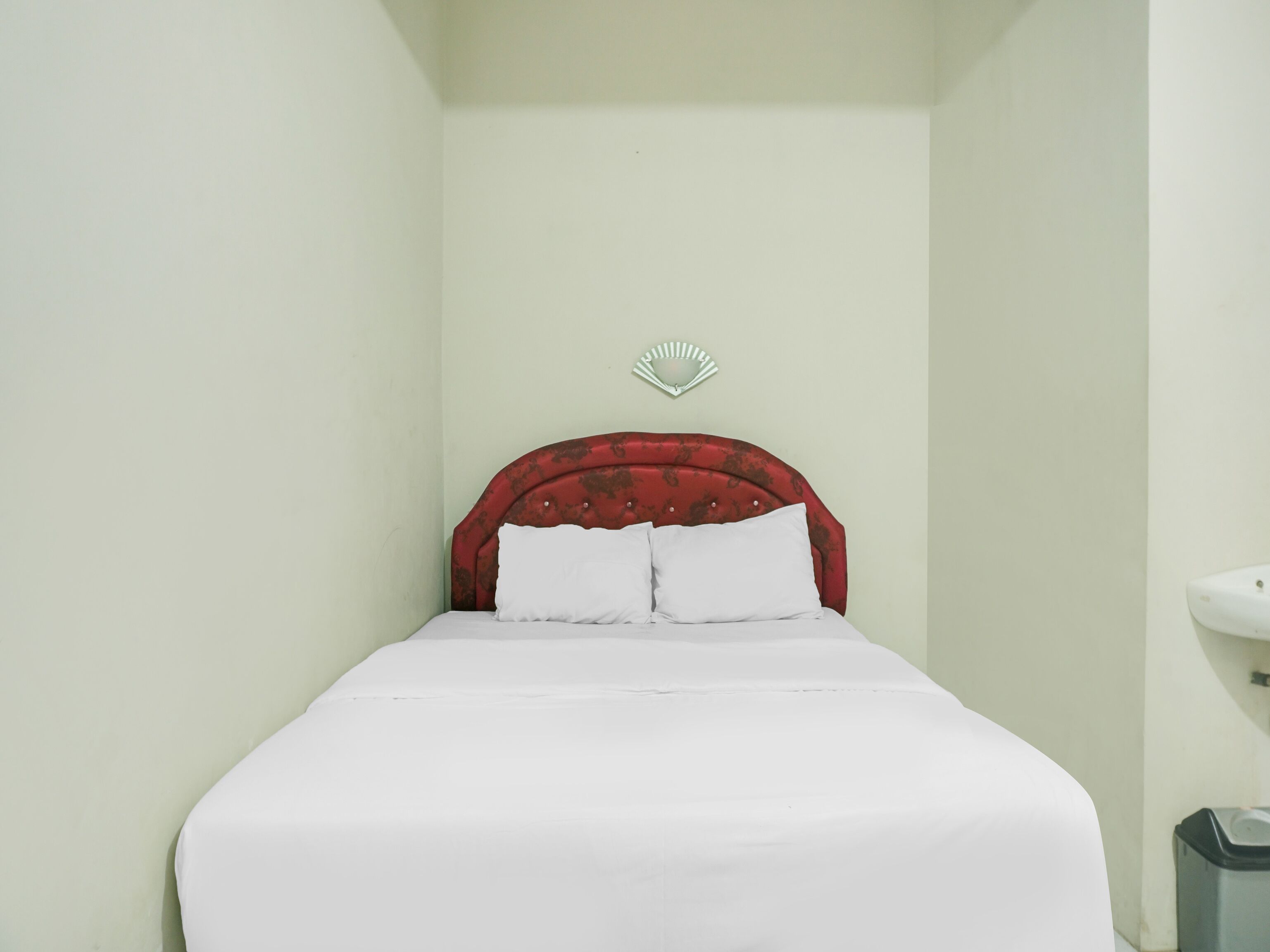 A small, single-bed room with a red headboard, white bedding, and a wall-mounted light fixture.