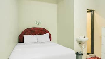 A neatly made bed with a red headboard and white bedding, a white sink, and a trash bin in a small bathroom.
