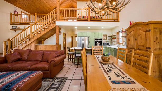 A spacious living area with a wooden staircase, a dining table with chairs, a sofa, and a chandelier with antlers. - Hummingbird Hacienda - Los Ojos Cabin w/ Brazos Cliff Views (Los Ojos)