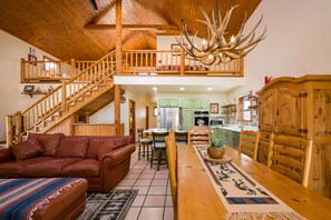 A spacious living area with a wooden staircase, a dining table with chairs, a sofa, and a chandelier with antlers. - Hummingbird Hacienda - Los Ojos Cabin w/ Brazos Cliff Views (Los Ojos)