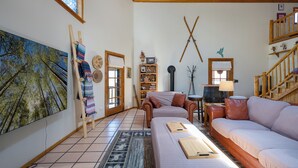 A cozy living room with a large painting, a fireplace, and a wooden staircase. - Hummingbird Hacienda - Los Ojos Cabin w/ Brazos Cliff Views (Los Ojos)