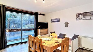 A dining area with a wooden table and chairs, a kitchen in the background, and a large window with a view of trees and a building. - Comfortable refurbished studio by the slopes, balcony, pool access (Le Corbier)