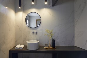 A modern bathroom with a round mirror, a white sink, and a vase with flowers. - Mayu Suites (Santorini)
