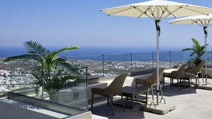 Terraza en la azotea con balaustradas de cristal, mobiliario exterior y sombrillas, con vistas a un pueblo costero.
