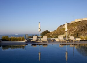 A rooftop pool with lounge chairs and umbrellas, overlooking a scenic view of a hill and the sea.