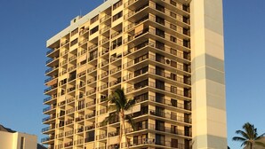 A high-rise hotel with multiple balconies, located near a sandy beach with palm trees.