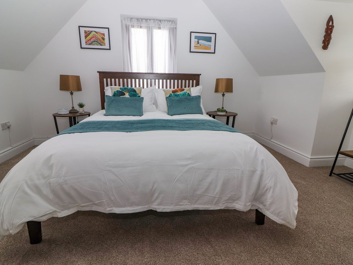 Una cama bien tendida en una habitación con techo inclinado, dos mesitas de noche con lámparas y dos cuadros enmarcados en la pared.