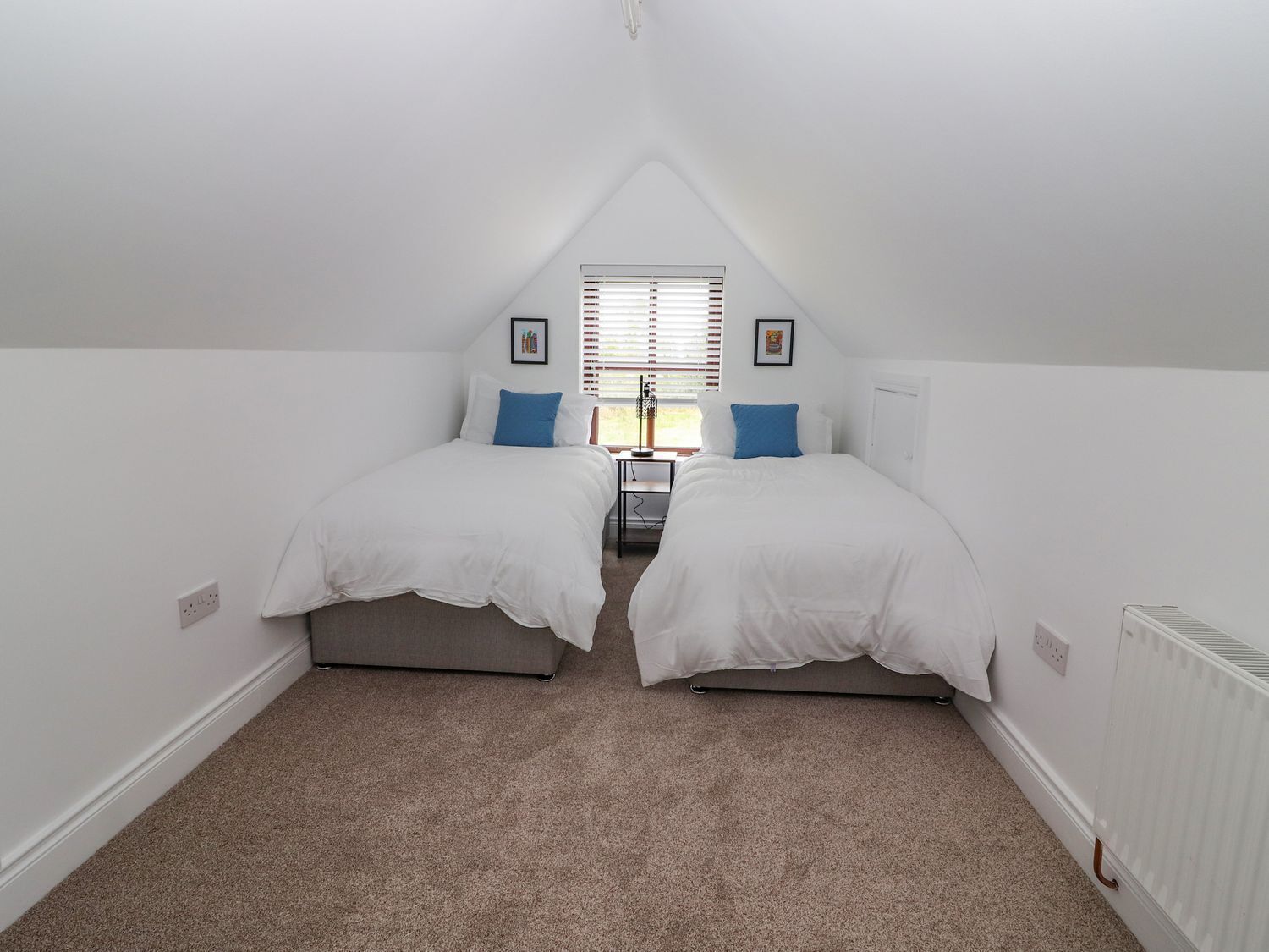 A small, white, attic-style bedroom with two beds, a radiator, and a window with blinds.