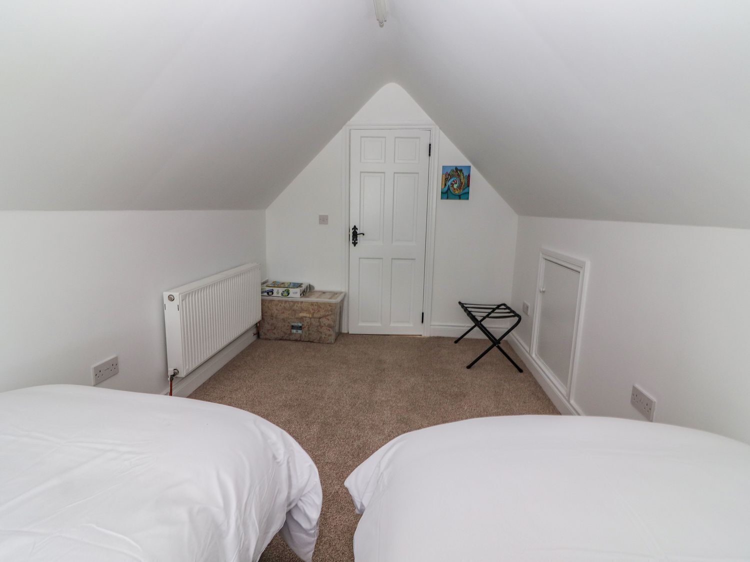 A small, white, attic-style bedroom with two single beds, a radiator, a door, and a small shelf with items.