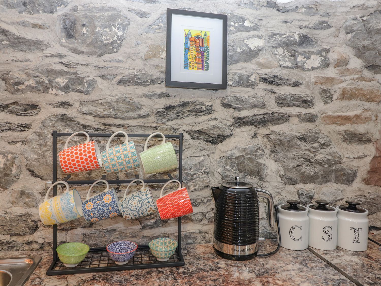 A kitchen with a stone wall, a framed cityscape, a mug rack, a kettle, and labeled storage containers.
