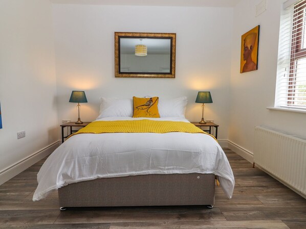 A neatly arranged bedroom with a bed, two bedside lamps, a framed mirror, and a window with blinds.